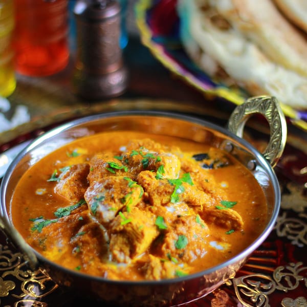 A copper bowl of Indian butter chicken, garnished with fresh cilantro, and naan bread in the background.
