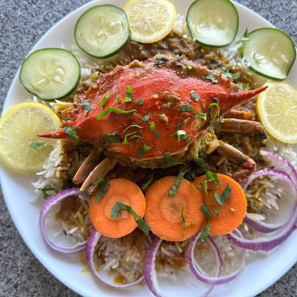 A whole red crab biryani, garnished with cucumber, lemon, carrots, and red onion on a white plate.